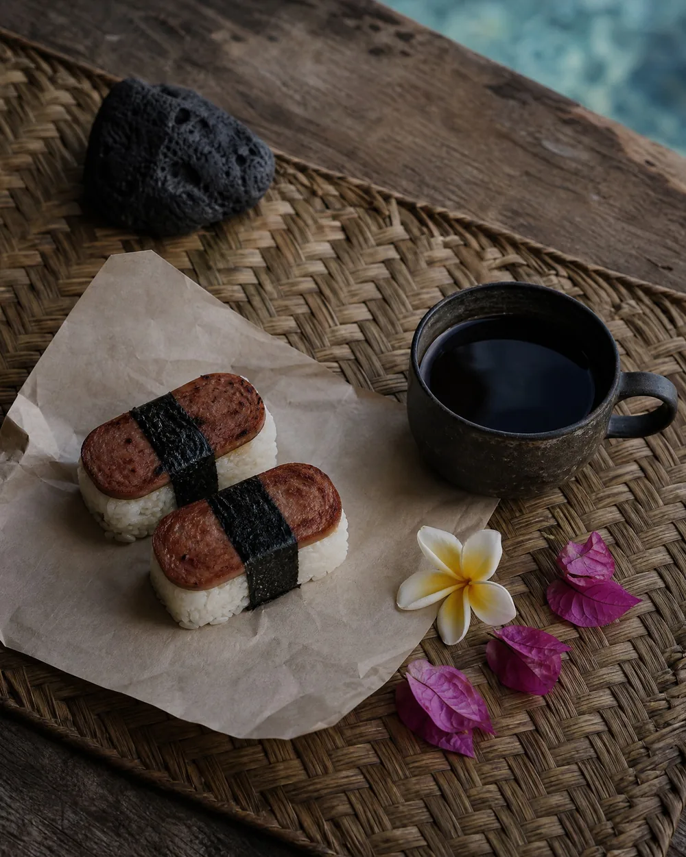Two Spam musubi on parchment beside a cup of black coffee, on a woven lauhala mat at a weathered table with the water beyond