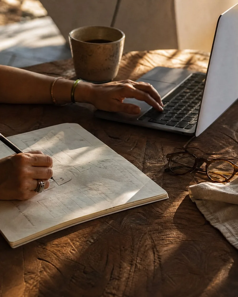 Hands at a laptop and a hand-drawn plan on a carved wood table, coffee and reading glasses beside them