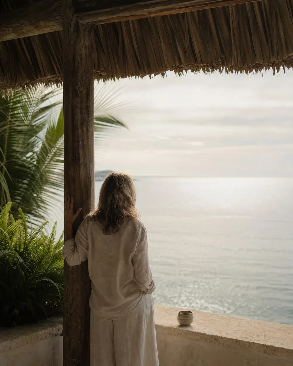 A woman standing at a timber post under a thatched roof, looking out over a bright calm sea