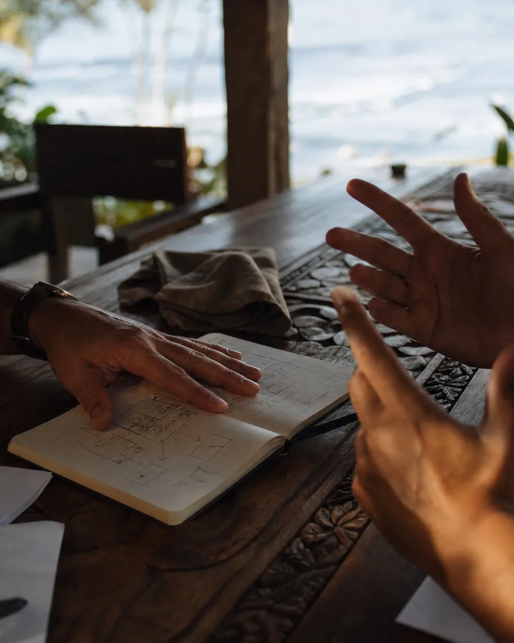 Two people talking across a teak table over an open notebook, a diagram sketched on the page, the ocean beyond